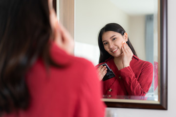 Beautiful Asian Woman wearing red dressed putting Powder puff looking in mirror in her bedroom at home. Makeup in morning getting ready before going to work.