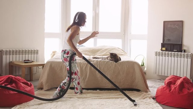 Young woman dancing with vacuum cleaner doing chores cleaning house having fun silly dance listening to music wearing headphones enjoying carefree weekend morning at home. 3840x2160