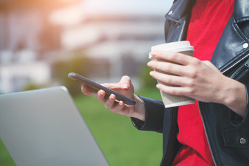 young woman holding and using smartphone and touching screen, drinking coffee and working on laptop outside