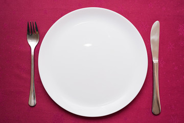 Empty white plate, fork and knife on maroon tablecloth. Elegant table setting. Top view, closeup
