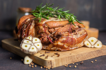 Baked pork brisket with twine on a wooden board with rosemary and spices and garlic on a dark background