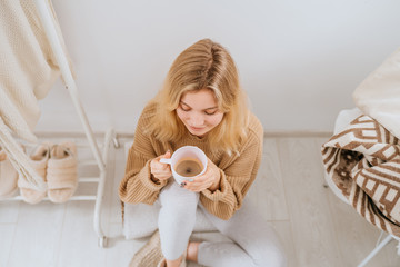 Young blond positive woman sitting in her light modern dressing room on the floor and drinks morning coffee. Top view. Fashion blog, website, social media hero header template
