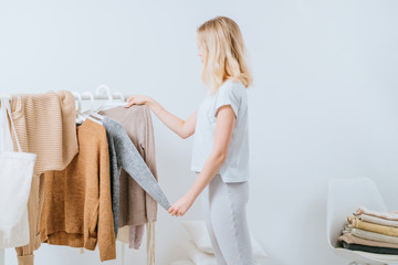 Young pretty girl blogger stands with a thoughtful expression on his face, having difficult choice not knowing what to put on on the background of clothes hanging on a hanger in the wardrobe.