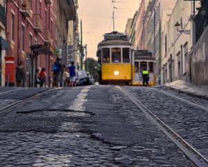 Fototapeta na wymiar Travel concept of Lisbon famous trams. Focusing on the cobbled streets with the trams in the foreground. In the background, tourists and trams out of focus, in the light of the sunset. Lisbon, Portuga