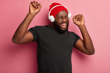 Feeling happy and relaxed. Joyful dark skinned man enjoys funny melody in headphones, has fun and entertainment, raises both hands, dressed casually, isolated on pink wall. Modern technology