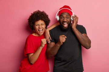 Youth lifestyle and party concept. Joyful dark skinned woman and man sing songs loudly, have overjoyed expressions, dance together, use headphones, isolated over rosy background. Music, technology