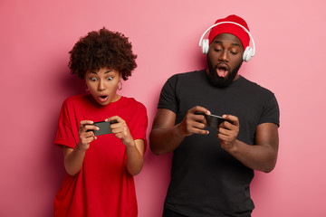 Addicted dark skinned girlfriend, boyfriend compete in video games, hold smartphones, focused at...
