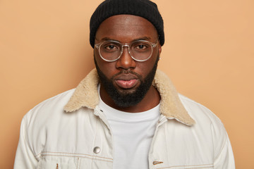 Cropped image of serious black man with full lips, thick beard, wears optical glasses, white shirt,...