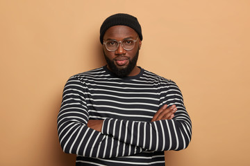 Self assured bearded male youngster stands with hands crossed, looks confidently at camera, has...