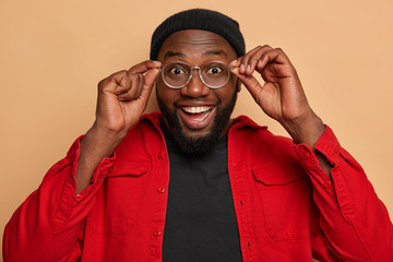 Portrait of attractive man with broad smile, keeps hands on rim of glasses, dressed in red shirt,...