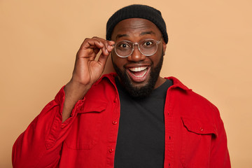 Headshot of cheerful dark skinned man keeps hand on rim of glasses, dressed in fashionable clothes, has broad smile, white perfect teeth, happy to hear best friend, meets someone. Emotions concept