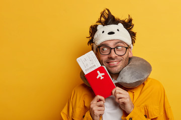 Happy man passenger holds passport and boarding pass ticket, has air travel flight, wears necessary items for sleeping in transport, enjoys traveling, looks aside with smile, isolated on yellow wall