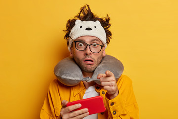 Impressed man points at you, holds cellphone horizontally, wears transparent glasses, neck pillow and shirt, traveles by blane, isolated over yellow background. People, technology, transport concept