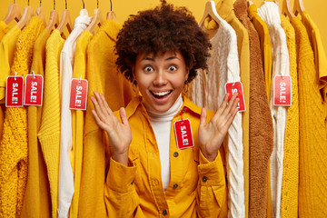 Time to refresh wardrobe. Positive curly woman spreads palms, tries on yellow shirt, reacts on sale...