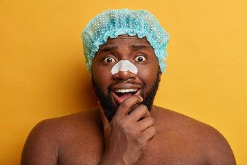 Black surprised bearded man holds chin, wears clear up strip on nose to remove blackheads, wears...