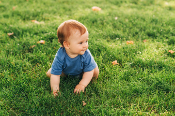 Adorable red haired baby boy crawling on fresh green grass in summer park