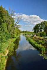 Ujście kanału wodnego do rzeki wśród drzew i chmur na niebie w letni dzień.
