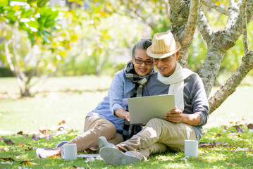 Relax Senior Asian couple  sitting under the tree  at park  using  laptop computer while holding coffee cup