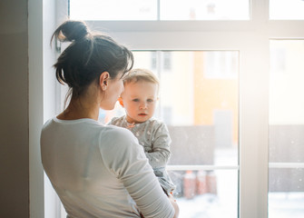 Young woman mom with baby girl on hands near window at home