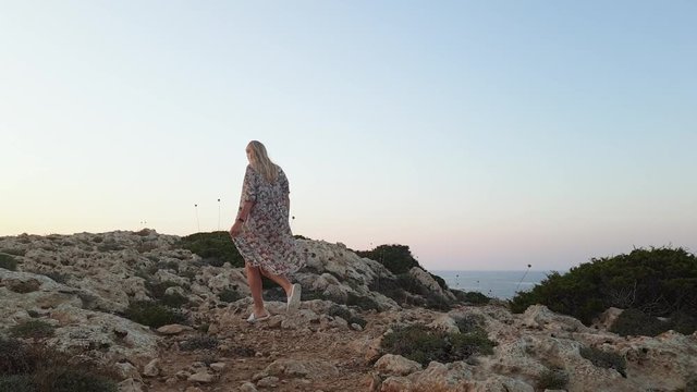 young slender woman with long blond hair in a bikini walks on the rocks at Cape Greco against the sea at sunset, pareo flutters in the wind, the concept of freedom and travel, slow motion