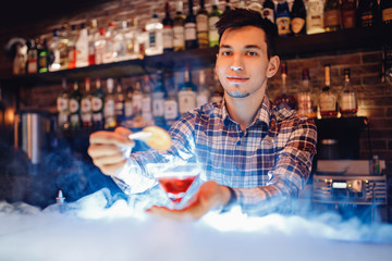 Barman is preparing red alcoholic cocktail with blue smoke in martini glass, decorated with orange