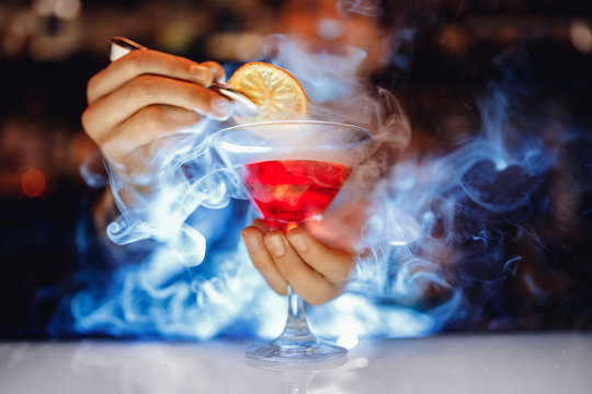 Barman is preparing red alcoholic cocktail with smoke in martini glass, decorated with orange