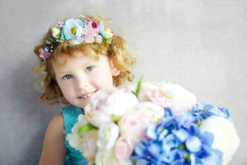Portrait of 5 years old curly blonde smiling girl in blue dress and wreath with big bouquet of white, blue and light rose artificial flowers. Waiting for spring small beautiful girl