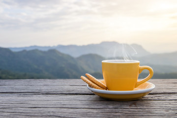 Eine weiße Tasse heiße Espresso-Kaffeetassen mit Keksen auf einem Holzboden mit Morgennebel und Bergen mit Sonnenlichthintergrund, Kaffeemorgen