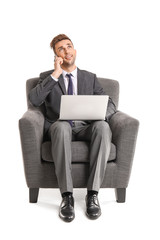 Handsome businessman with laptop talking by mobile phone while sitting in armchair on white background
