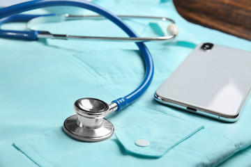 Stethoscope, mobile phone and doctor's uniform on table, closeup