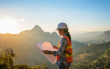 Engineer Asian man with long hair wearing a white helmet looking at the blueprint with sunset on...