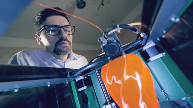 A man watches printing machine creating an orange vase. Professional industrial designer creates 3d printed product in a designing agency.