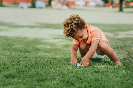 Cute african toddler boy playing outside with toy cars in the park on green grass, wearing orange polo t-shirt