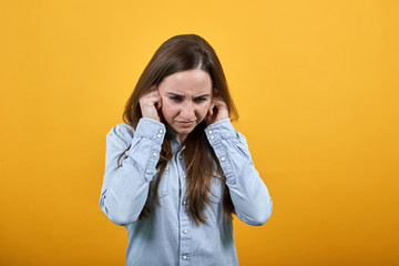 Tired woman in denim blue shirt isolated on orange background in studio closed ears with fingers, hate everything. People emotions, lifestyle concept.