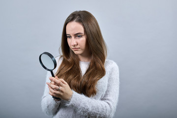 Attractive young caucasian woman looking on magnificent glass, wearing fashion blue sweater isolated on gray background in studio. People emotions, lifestyle concept.