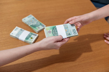 Woman hand giving pile of Polish money to woman hand. Wooden table background with piles of money. Polish money banknotes