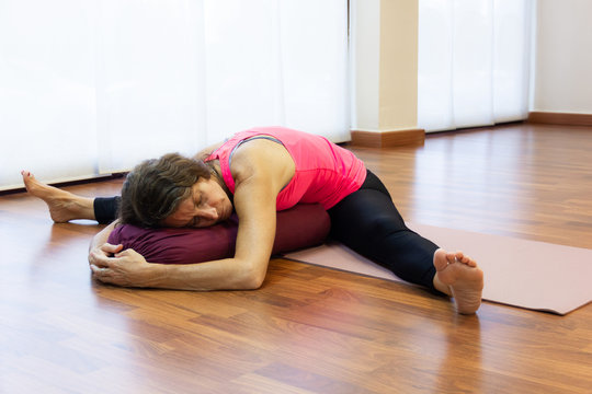 Middle aged woman in dragonfly pose lying forward on bolster. Young lady with open legs in straddle asana at bright studio. Yin yoga class concept