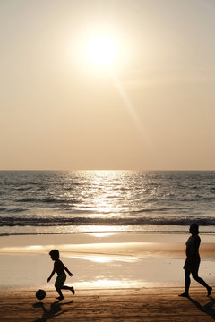 Silhouette of an unrecognizable small child kicking a footaball followed by his mother on a golden sandy beach at sunset infront of a calm sea