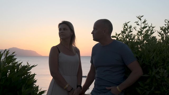A woman and a man of 45 years old are standing near the fence and looking at the rising sun beyond the horizon. The husband and wife are on vacation at the resort.