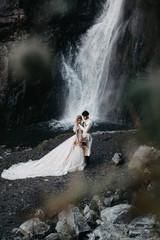 noise effect, selective focus: incredibly enamored brides hugging, kissing and posing for a photo on the incredible rocky mountains background with a big waterfall