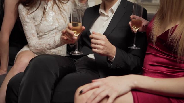 Unrecognizable womanizer young man flirting with three beautiful women sitting with him on sofa and stroking seductively his leg and hand at party
