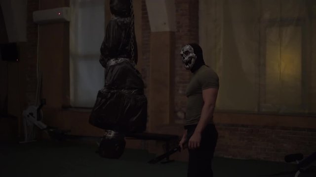 Maniac in silver skull mask, examines his victim, who weighs in a black bag