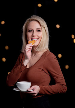 Blonde girl with cup of coffee and gingerbread cookie on background with fairy lights