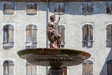Sculpture of the fountain and the wall with many windows closed by shutters in the French village...