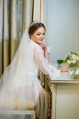 The bride in a white coat and veil sits at the makeup table