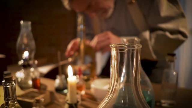 A mature white bearded scientist from the 18th century in his laboratory makes notes about observations he made through his microscope.