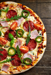 Pizza with ham, salami, jalapeno pepper, cherry tomatoes, cheese and rocket. Rustic wooden background. Top view.