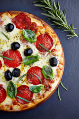 Pizza with salami, mozzarella cheese, black olives, and fresh herbs on black stone background. Top view. 