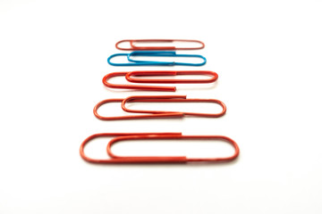 A blue paper clip amongst red paper clips stands out from the rest shot against a white background.