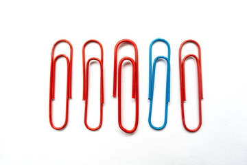 A blue paper clip amongst red paper clips stands out from the rest shot against a white background.